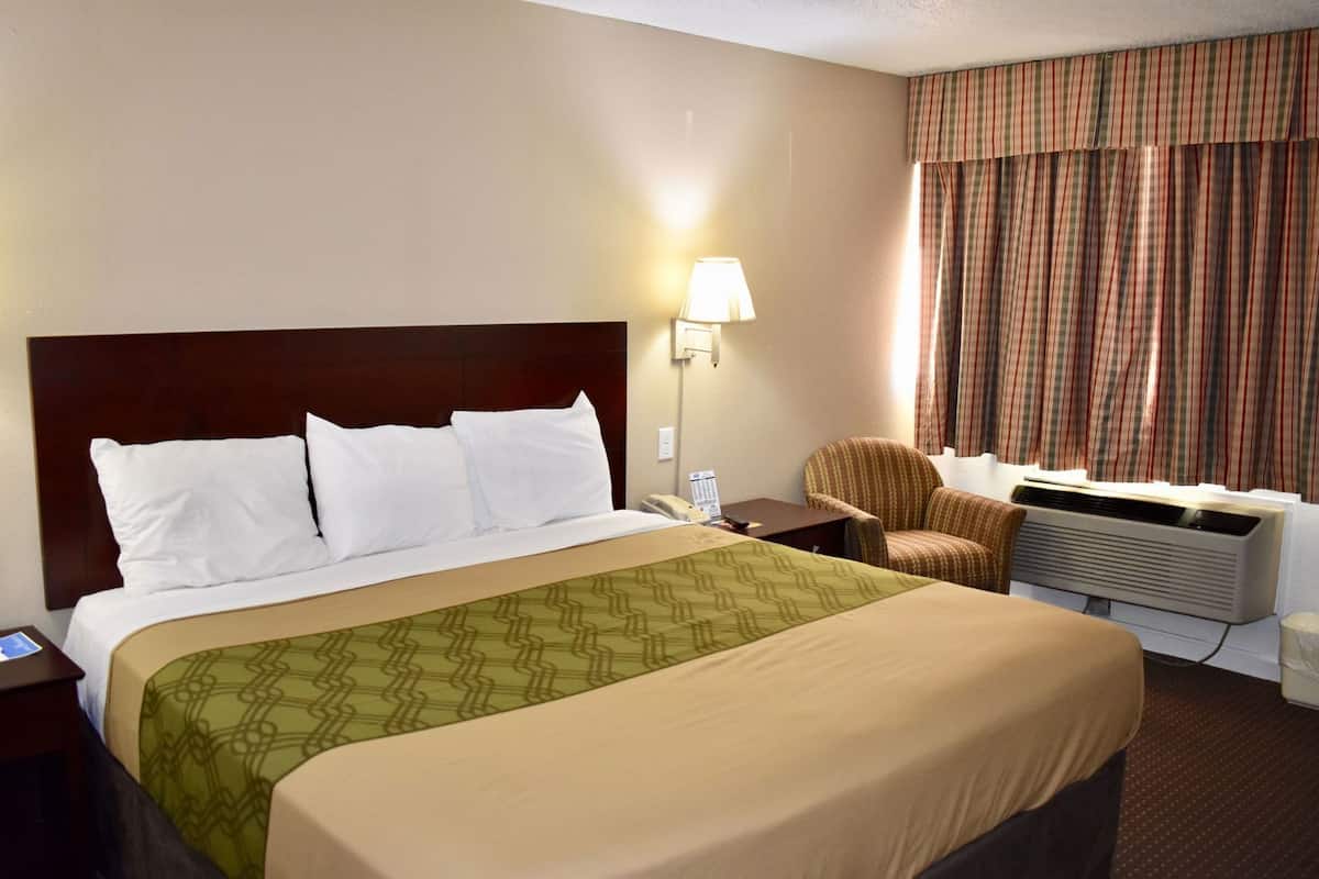 A hotel room with a bed, a chair, an air conditioning unit, and a lamp.