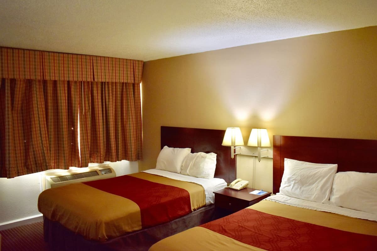 A hotel room with two beds, a bedside table with a lamp, a phone, and a window with curtains.