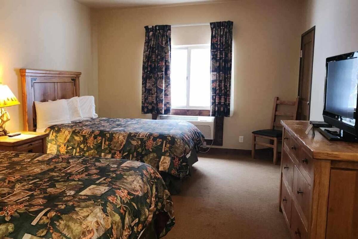 Quarto de hotel com duas camas, televisão, escrivaninha com gavetas, uma cadeira e janela com cortinas.