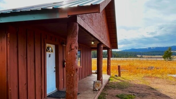 Una cabaña de madera con un porche, una puerta con el número 18 y vistas a un campo y montañas al fondo.