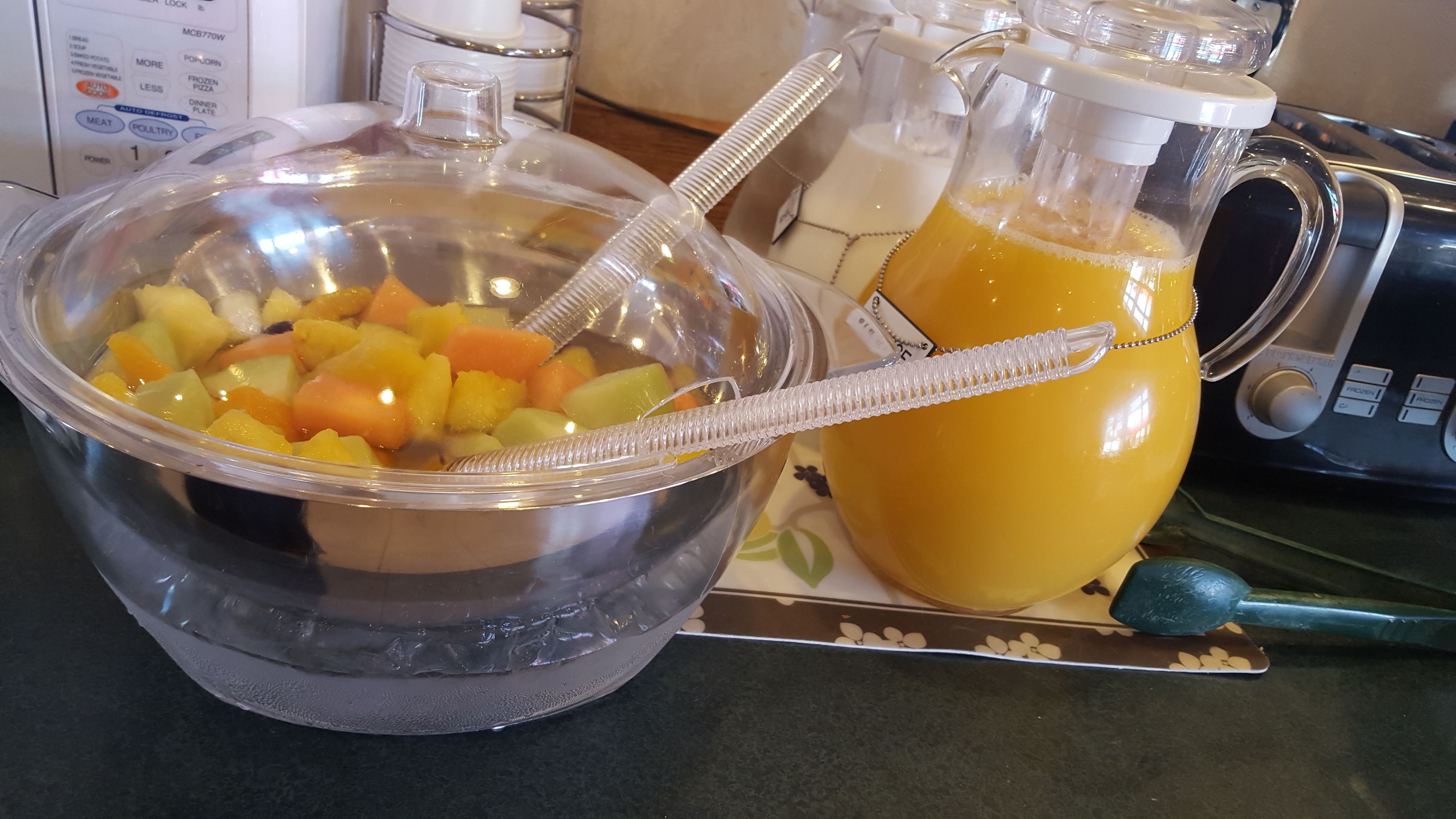 Una encimera de cocina con un frutero con frutas cortadas, una jarra de zumo de naranja y una licuadora.