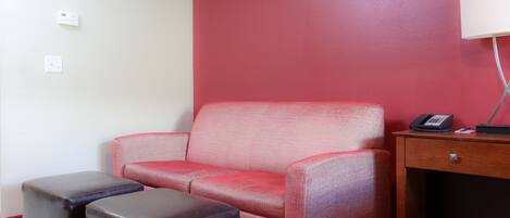 Ein rotes, gepolstertes Sofa, zwei schwarze Hocker, ein Nachttisch aus Holz mit einer Lampe und ein Telefon.