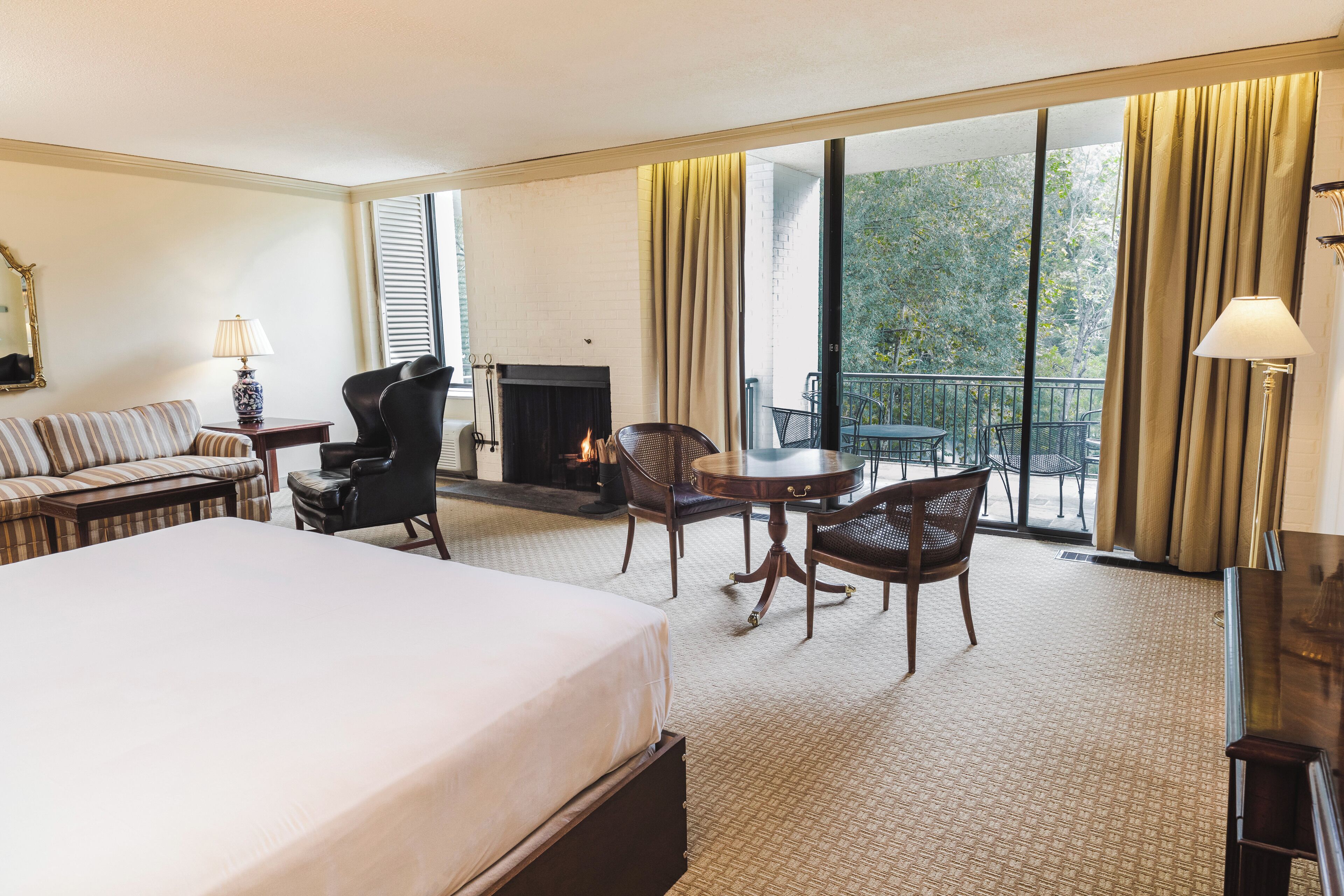 Habitación de hotel con una cama grande, un sofá, una chimenea, una mesa de comedor con sillas y un balcón con cortinas.