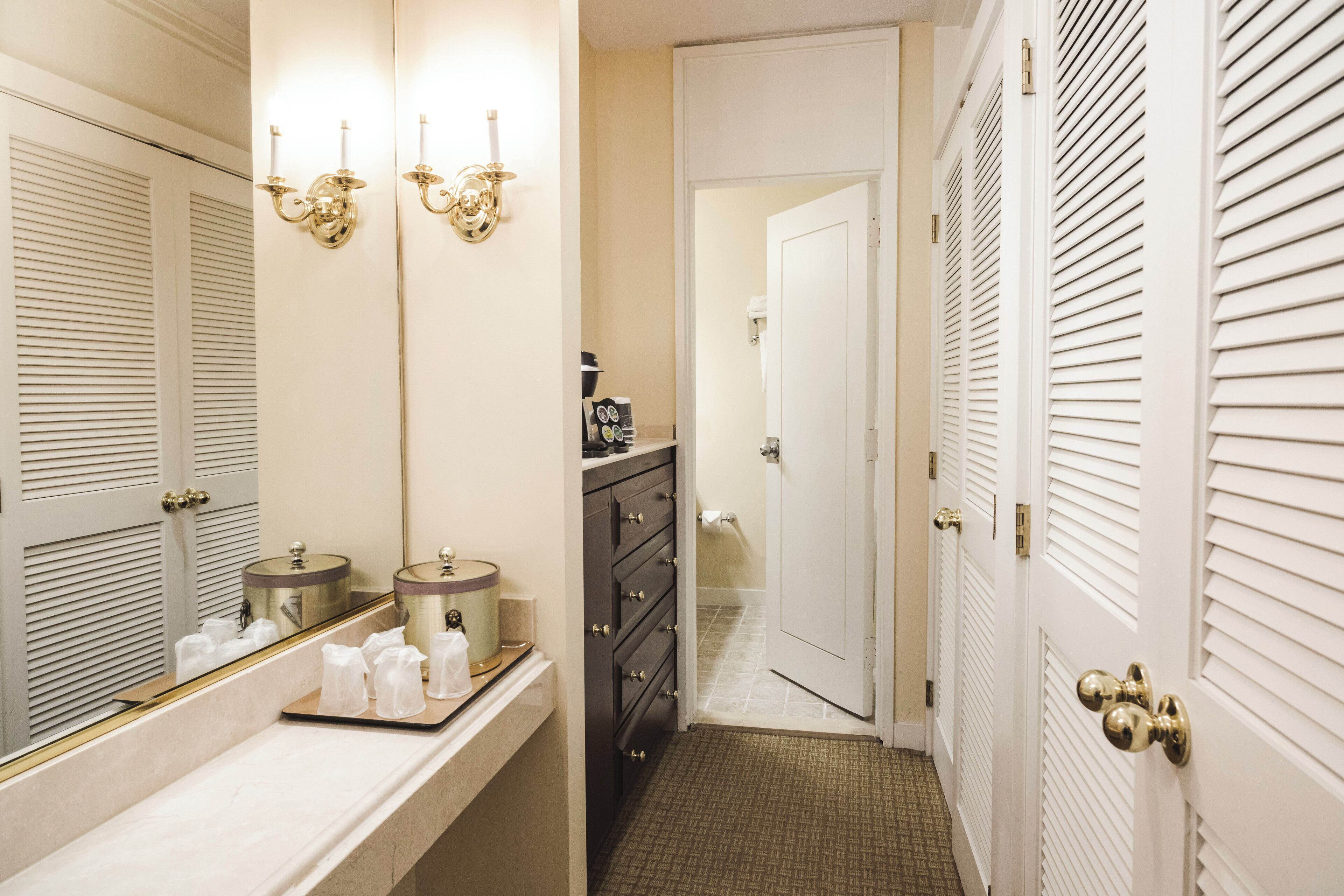 A bathroom with a vanity, two wall sconces, a mirror, and a door leading to another room.
