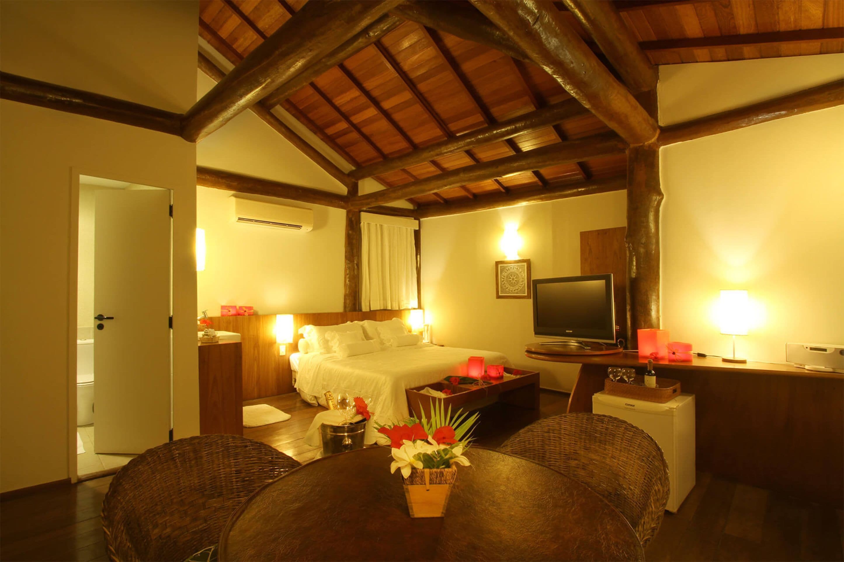 A hotel room with a wooden ceiling, a bed with white linens, a television, a small table with a vase of flowers, and a desk with a lamp.