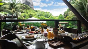 A breakfast table set with various dishes, drinks, and a view of lush greenery and a mountain in the background.