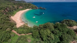 An aerial view of a coastal area with a sandy beach, clear turquoise waters, and lush green hills.
