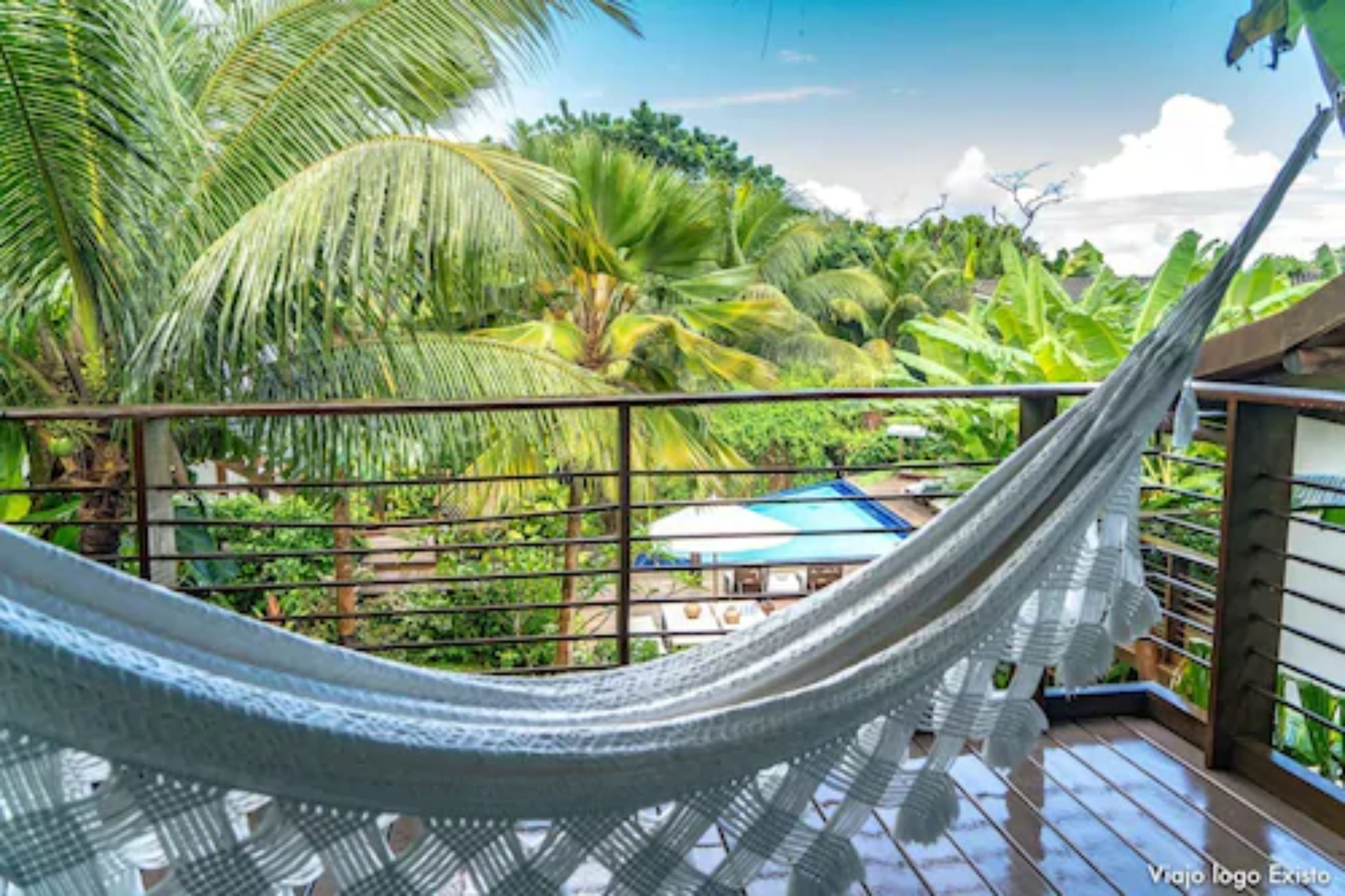 A hammock on a balcony with a view of lush greenery and a building with a blue roof.