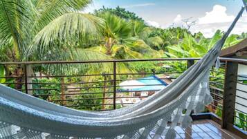 A hammock on a balcony with a view of lush greenery and a building with a blue roof.