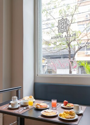 A breakfast table with various dishes and drinks, a window with a view of the street and a building with a sign. - Plaza Spania (Rio de Janeiro)