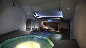 Un área de piscina interior moderna con un jacuzzi, un espacio de descanso y una cocina al fondo.