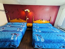 Una habitación con dos camas individuales, cada una con colcha azul estampada con delfines. Las camas tienen cabeceros de madera y están ubicadas frente a una pared roja. Hay una mesita de noche con una lámpara entre las camas.