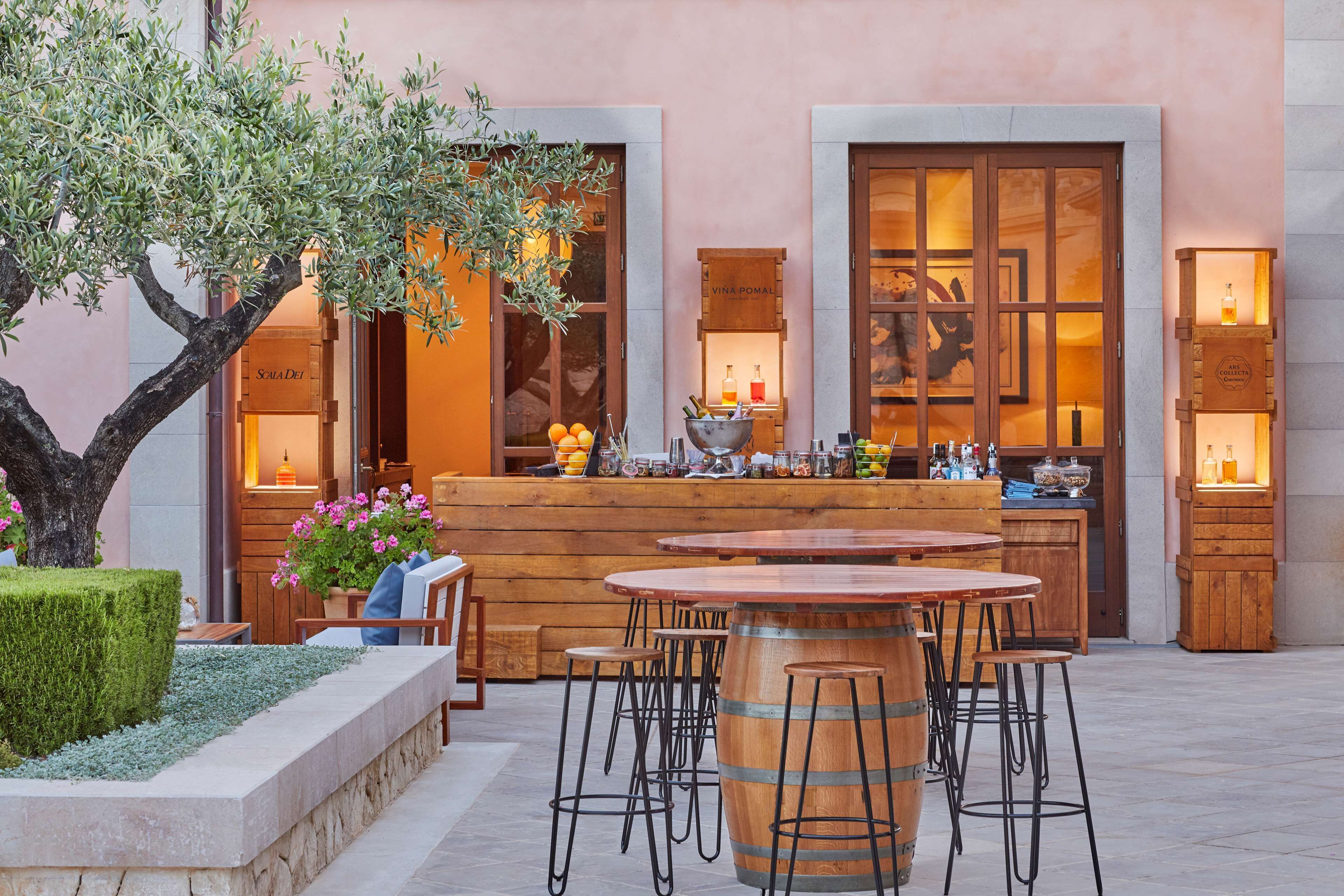 Un espace bar extérieur doté de mobilier en bois et d’un grand tonneau servant de pièce maîtresse, entouré d’un jardin bien entretenu et d’un bâtiment aux grandes fenêtres dont l’intérieur est chaleureux et accueillant.