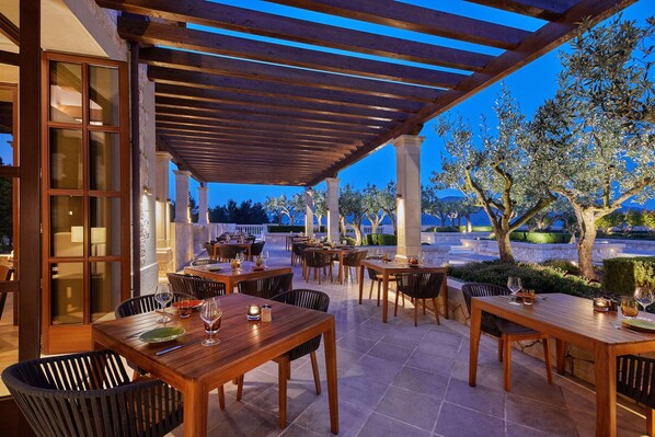 Un área de comedor al aire libre con mesas y sillas de madera, bajo una pérgola, rodeada de olivos y un muro de piedra.