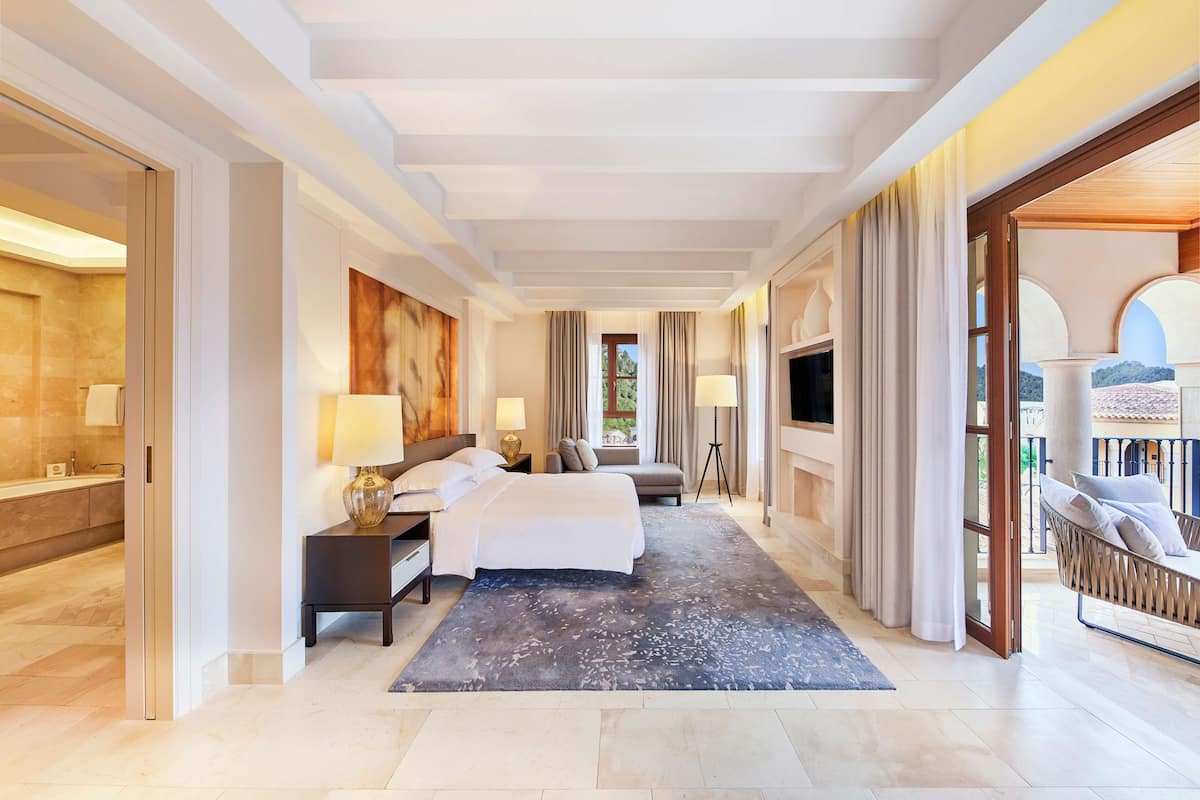 A spacious hotel room with a large bed, a sitting area, a TV, and a balcony with a view.