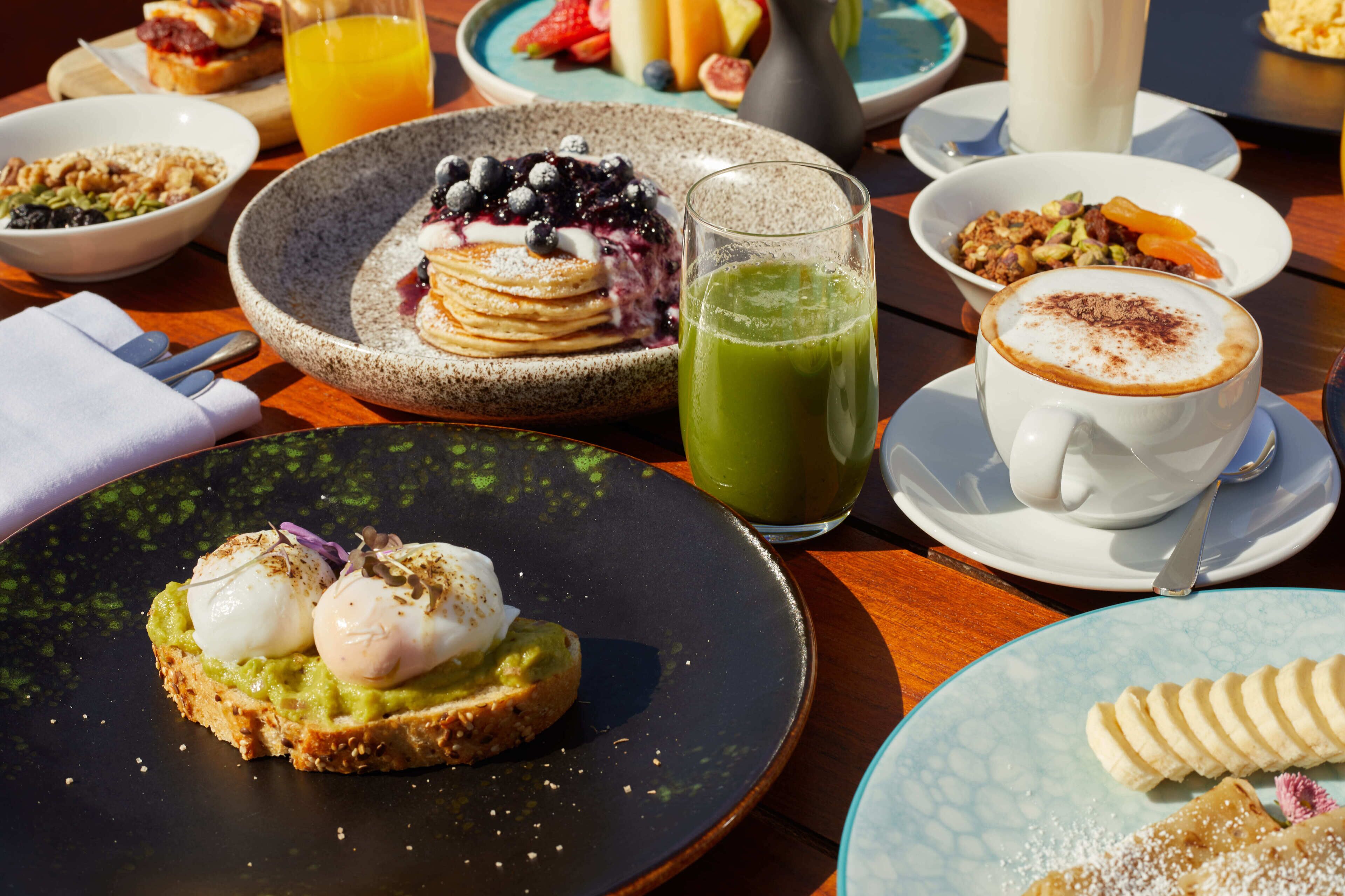 Una mesa de desayuno con diferentes platos, como panqueques, un batido verde y una taza de café.