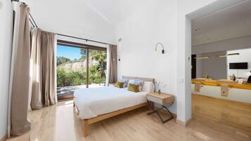 A modern bedroom with a large bed, a bedside table, a mirror, and a view of greenery outside.