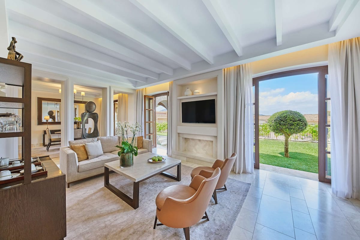 A modern living room with a sofa, coffee table, and chairs, a flat-screen TV, and a large window with a view of a garden.