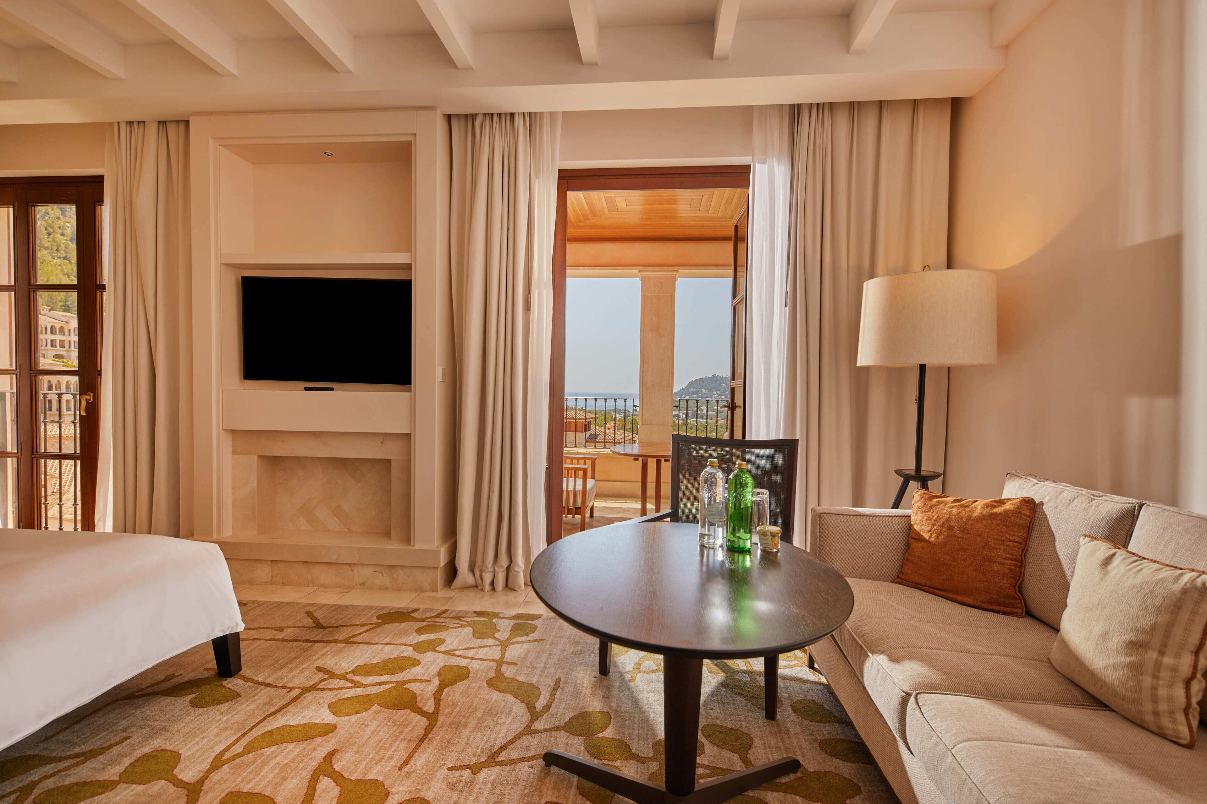 A hotel room with a bed, a sofa, a TV, a table with bottles, and a view of a building through the window.