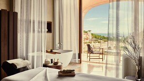 A modern bedroom with a large window, a bathtub, a chair, and a table.