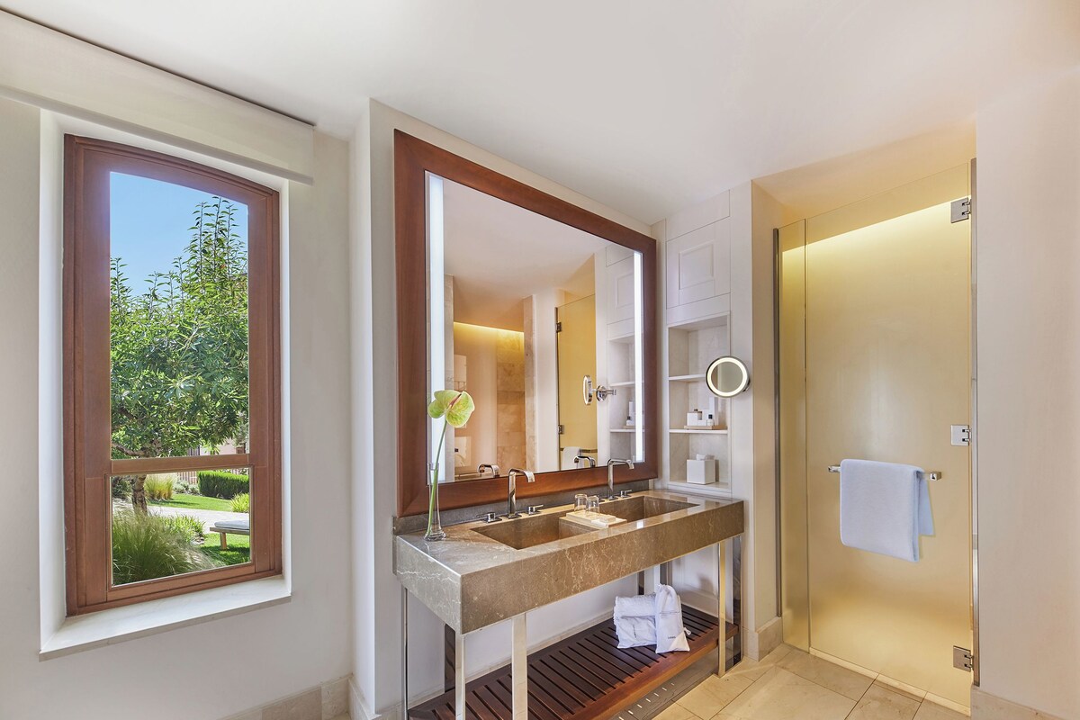 Un baño con lavabo doble, un espejo grande, zona de ducha y vistas a zonas verdes a través de una ventana.