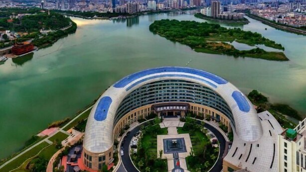 A large, modern building with a curved roof, surrounded by a landscaped area and a river.