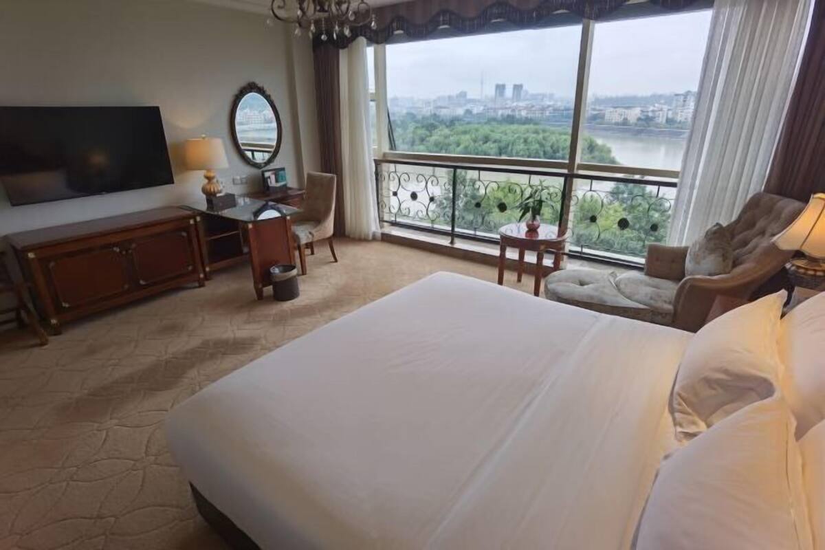 Ein Hotelzimmer mit einem großen Bett, einem Fernseher, einem Schreibtisch mit Stuhl, einem kleinen Tisch mit Pflanze und einem Balkon mit Blick auf Stadt und Fluss.