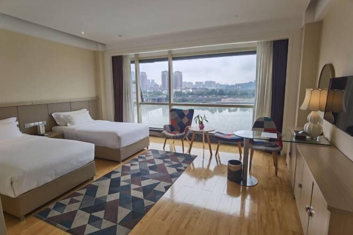 Ein Hotelzimmer mit zwei Betten, einem großen Fenster mit Blick auf die Stadt, einem kleinen Tisch mit Stühlen und einer Lampe.