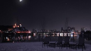 A nighttime view of a cityscape with a lake, chairs, and a partially snow-covered ground.