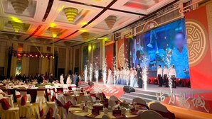 A banquet hall with round tables, red sashes, and a stage with a large screen.