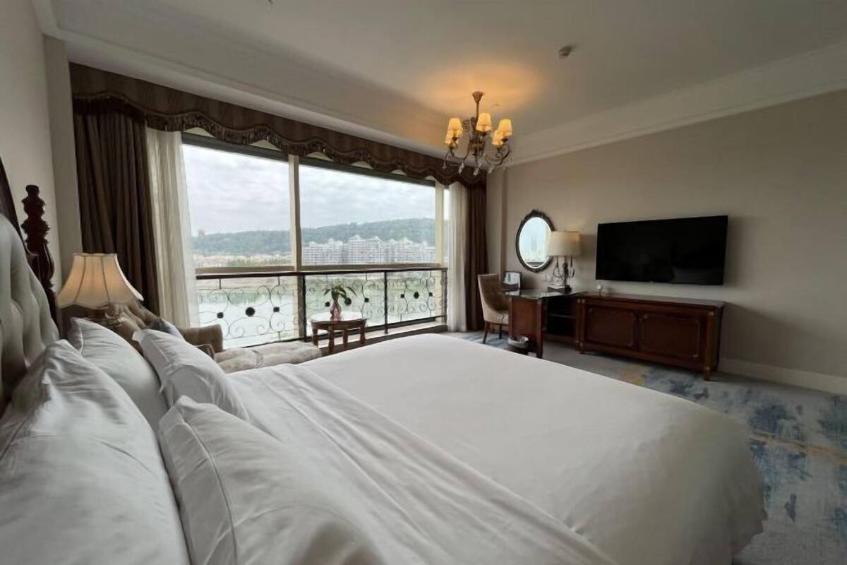 Ein Hotelzimmer mit einem großen Bett, einem Schreibtisch mit Stuhl, einem Fernseher und Blick auf die Stadt durch große Fenster.