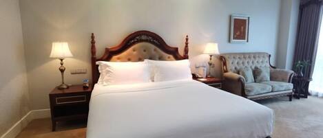 A large bed with a wooden headboard, two bedside tables with lamps, a tufted armchair, and a framed picture on the wall.