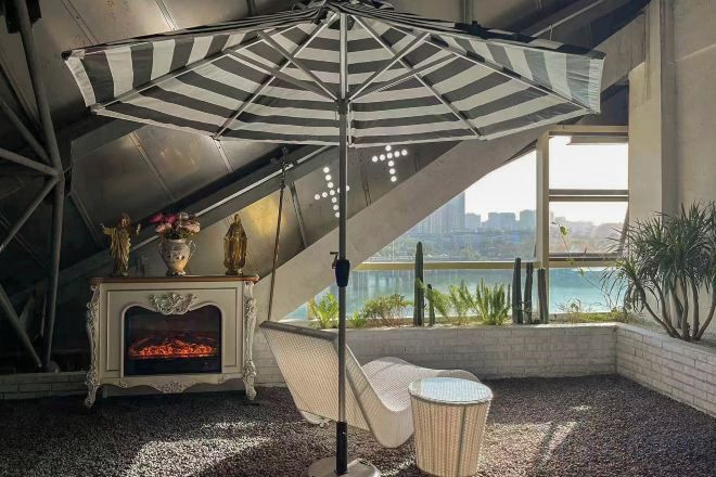 A room with a sloped ceiling, a hammock, a fireplace, a large window with a view, and a striped umbrella.