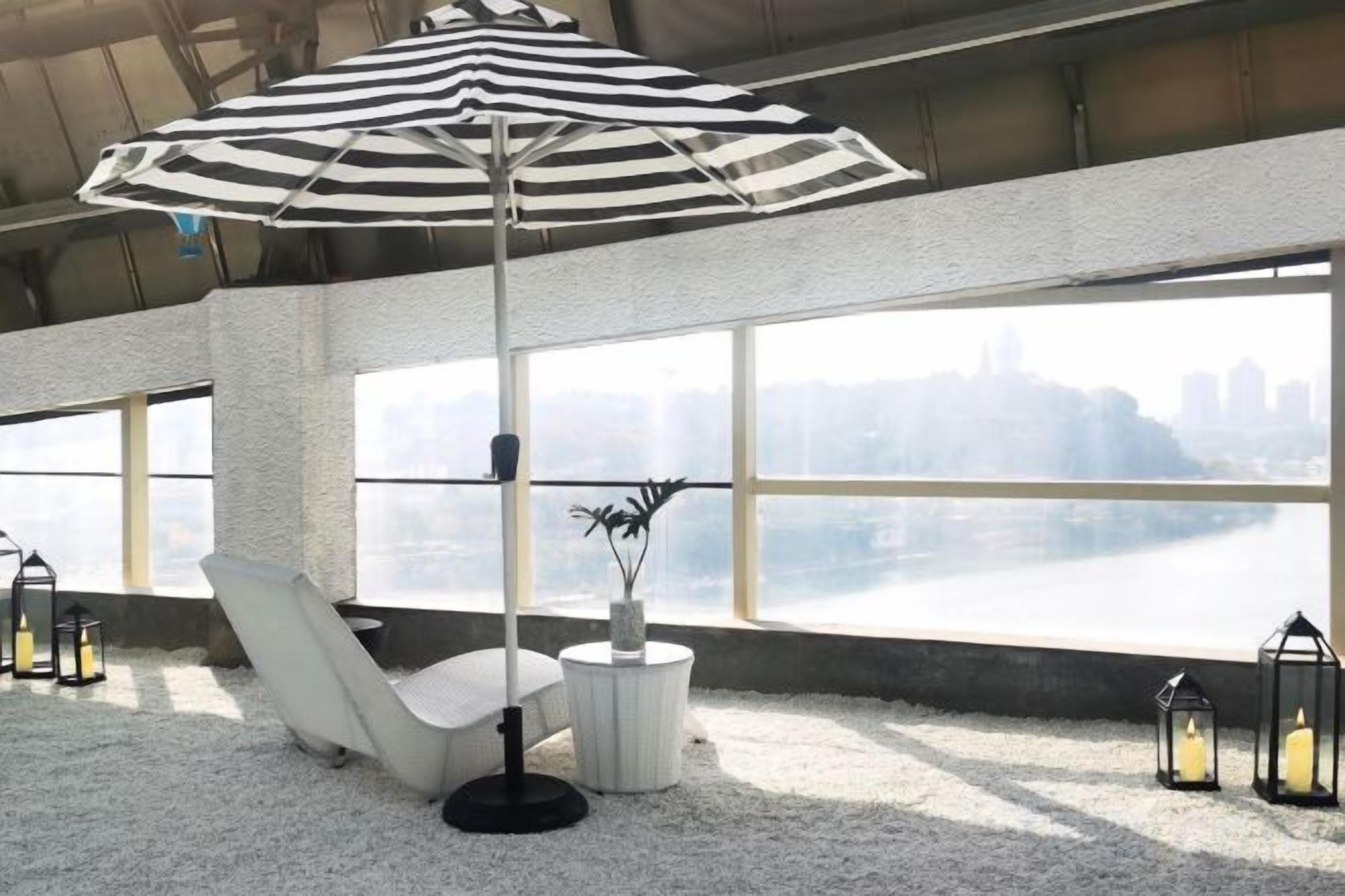 A lounge area with a white chair, a striped umbrella, and a view of a cityscape through large windows.