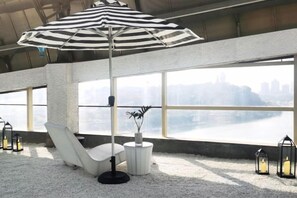 Un espace détente avec un fauteuil blanc, un parasol à rayures et une vue sur le paysage urbain, visible à travers de grandes fenêtres.