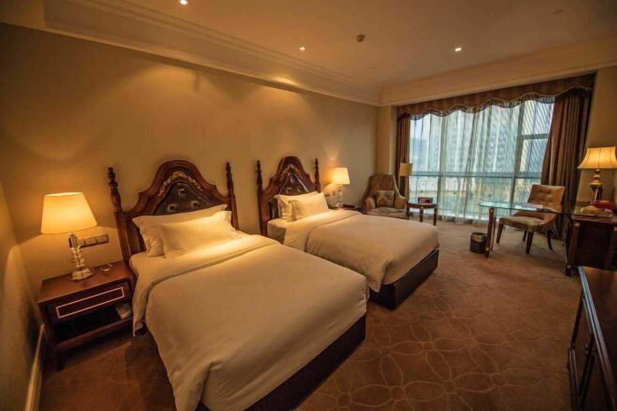Ein Hotelzimmer mit zwei Betten, einem großen Fenster mit Vorhängen, einem Sessel, einem Schreibtisch und einer Lampe.