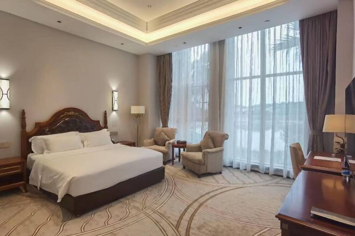Ein Hotelzimmer mit einem großen Bett, zwei Sesseln, einem Schreibtisch und einem großen Fenster mit Vorhängen.