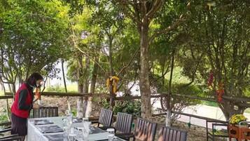 Un espacio de comedor al aire libre con una larga mesa dispuesta para invitados, rodeado de árboles y una valla de madera.