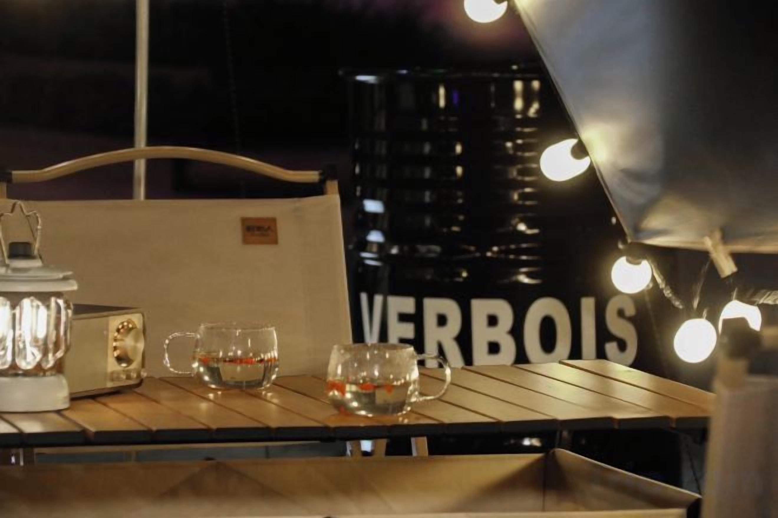 A table with a white lamp, two glasses, and a bottle, set against a backdrop with a sign reading 'VERBOIS'.