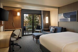 Ein Hotelzimmer mit Bett, Schreibtisch, Stuhl, Sofa, Fernseher und einem Balkon mit Sitzgelegenheit.