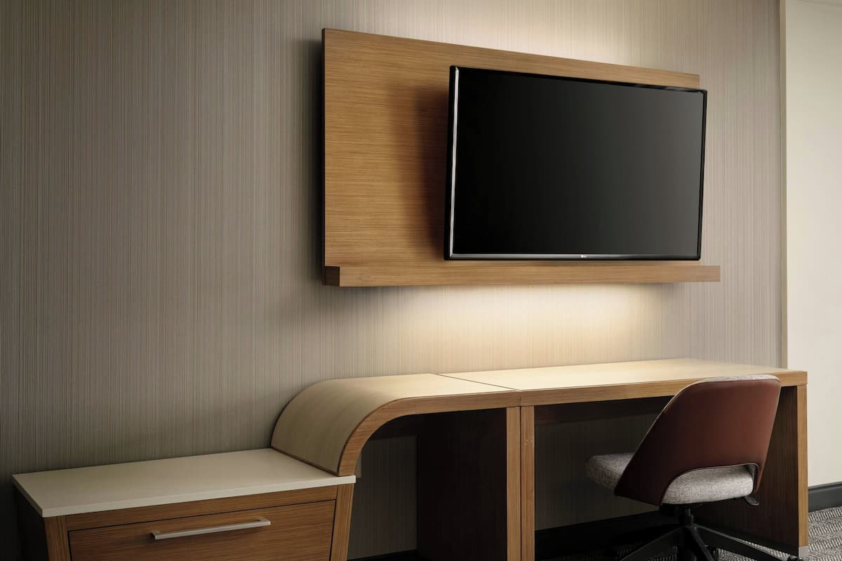 Une chambre d’hôtel moderne dotée d’un téléviseur à écran plat fixé au mur, d’un bureau en bois avec une chaise rembourrée et d’un tiroir intégré.