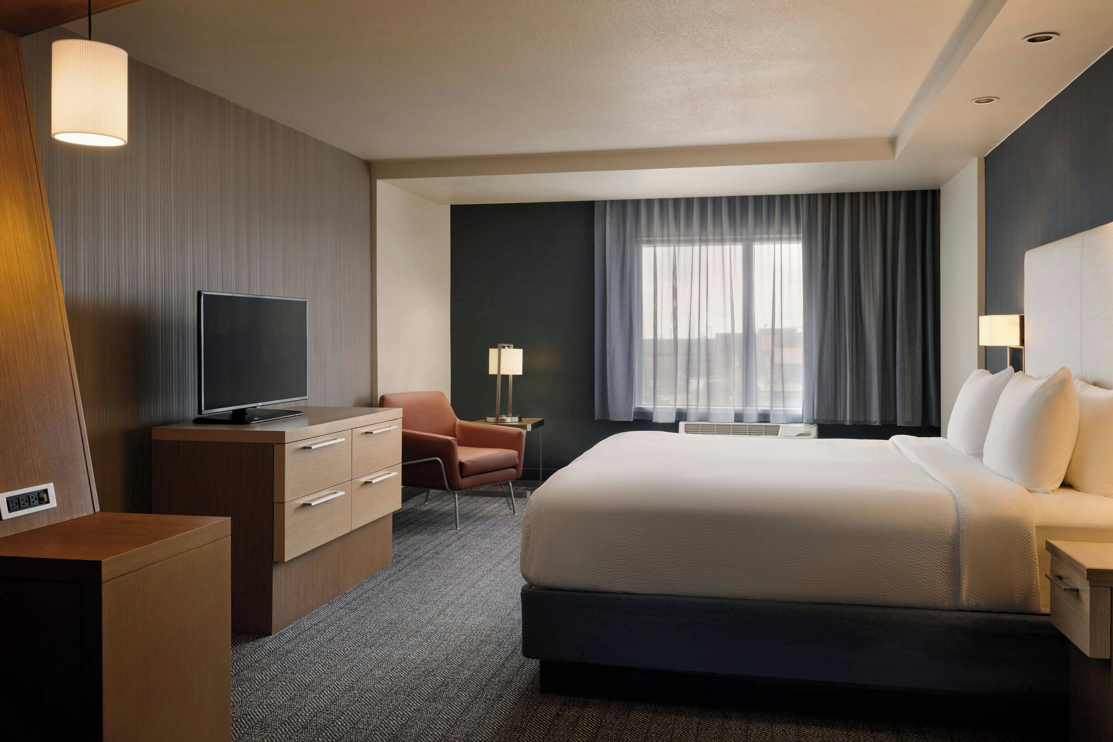 A hotel room with a bed, a desk with a television, a chair, a lamp, and a window with curtains.