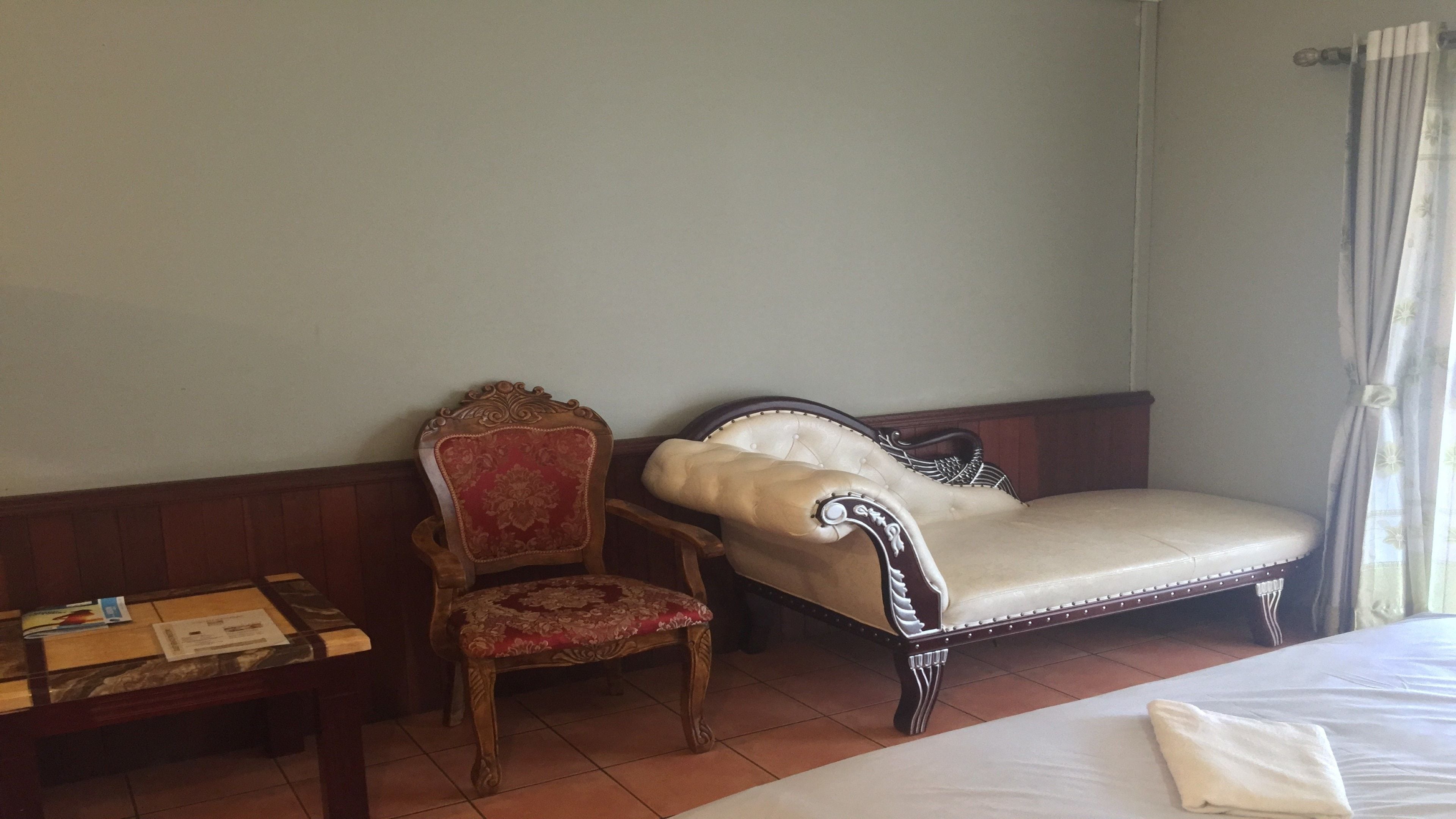 A hotel room with a bed, a wooden chair with a red cushion, a sofa with a white cushion, and a small table with a book and a piece of paper.