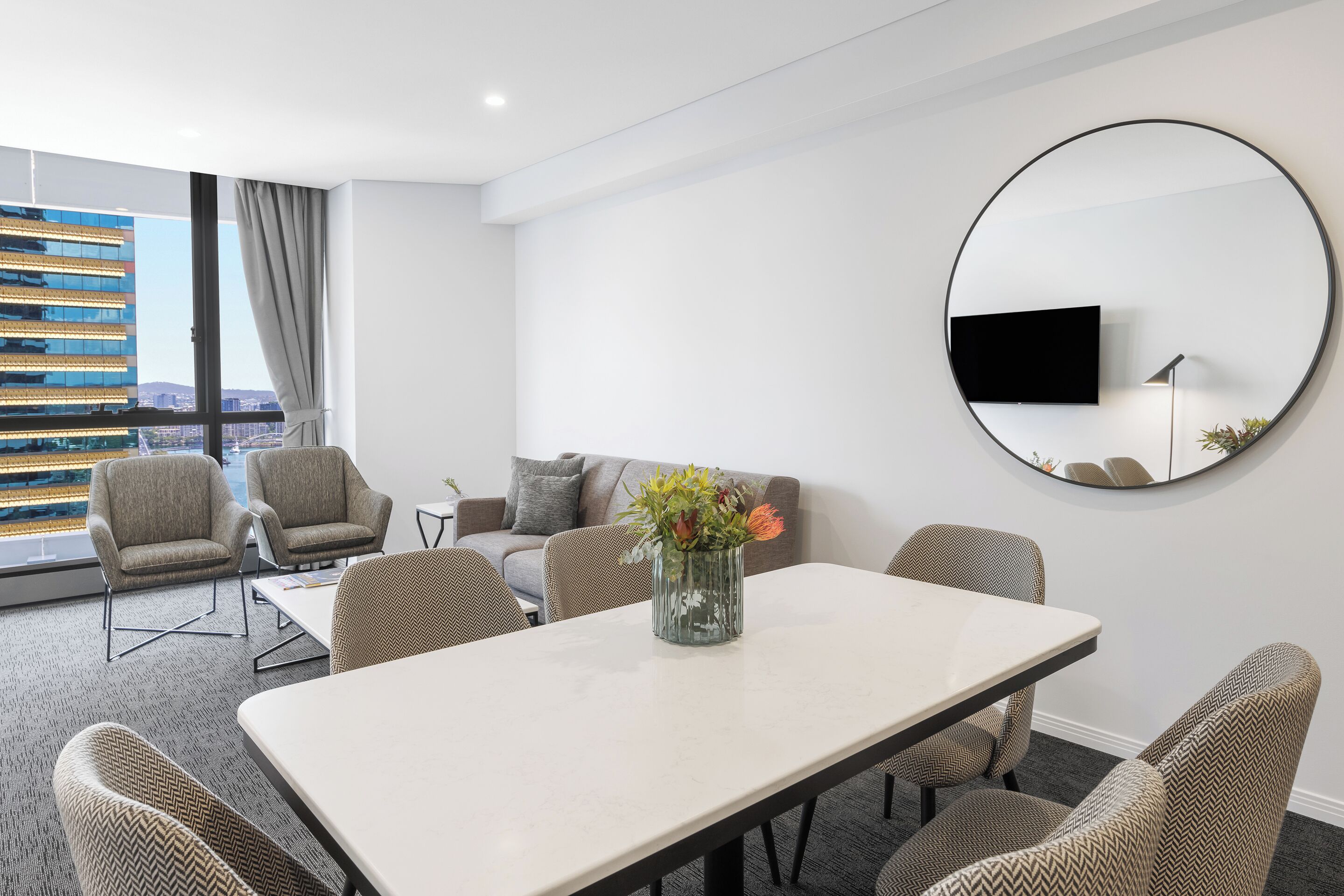 A modern hotel room with a dining area, a sofa, and a large window with a city view.