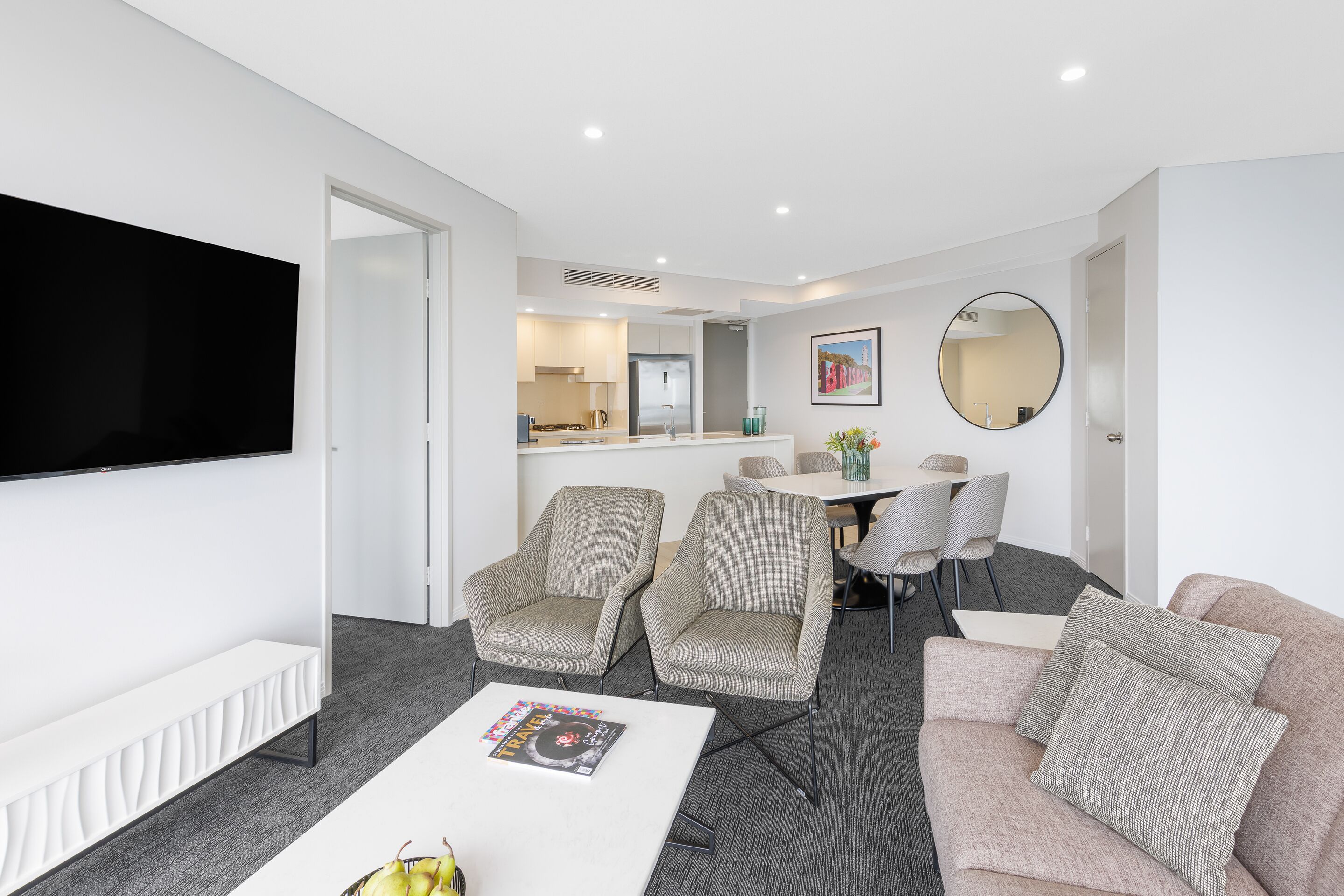 A modern living room with a flat-screen TV, two armchairs, a sofa, and a coffee table. There is a dining area with a table and chairs in the background.