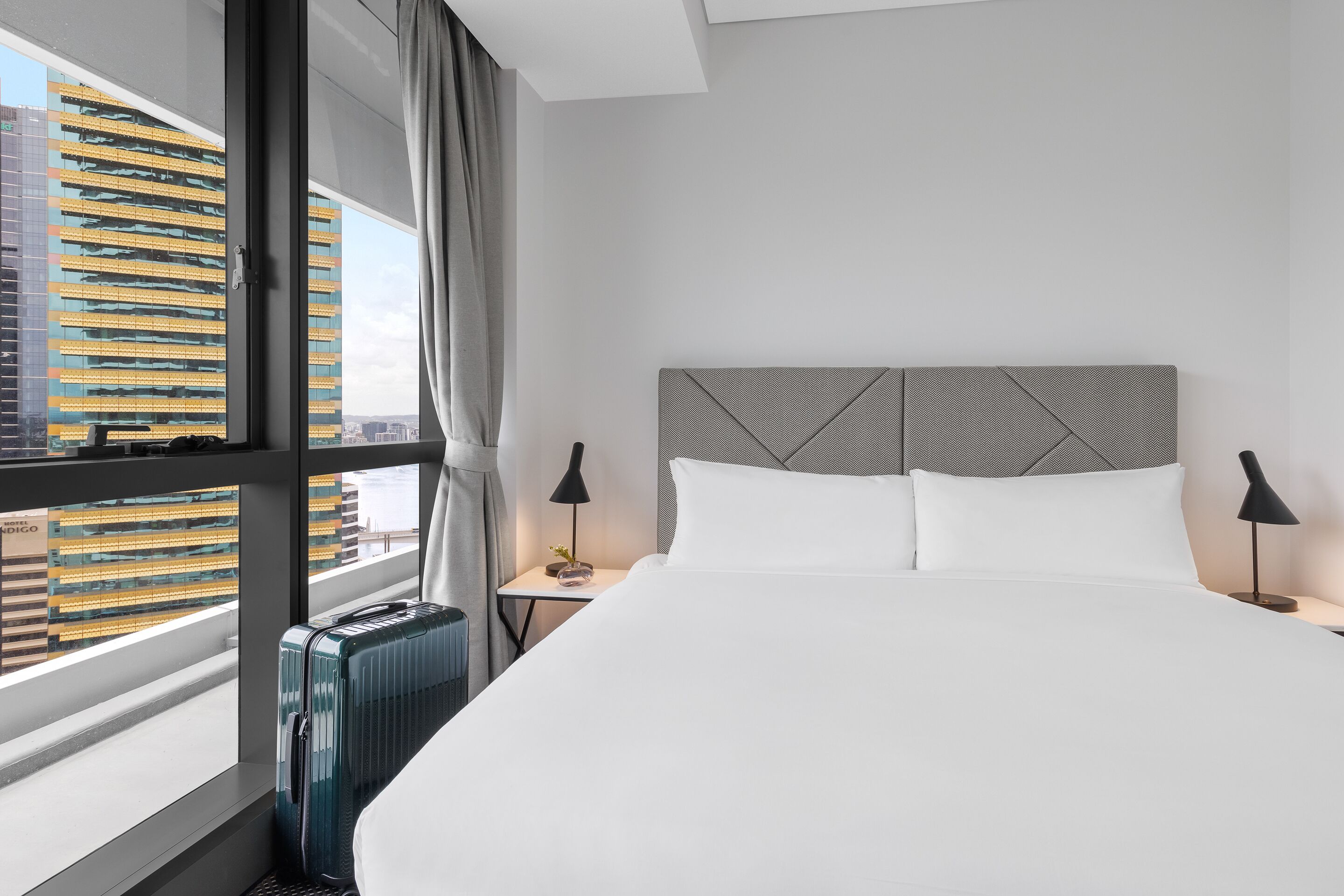 Une chambre d’hôtel moderne avec un grand lit, une table de chevet, une lampe et une vue sur le paysage urbain, visible à travers une grande fenêtre.