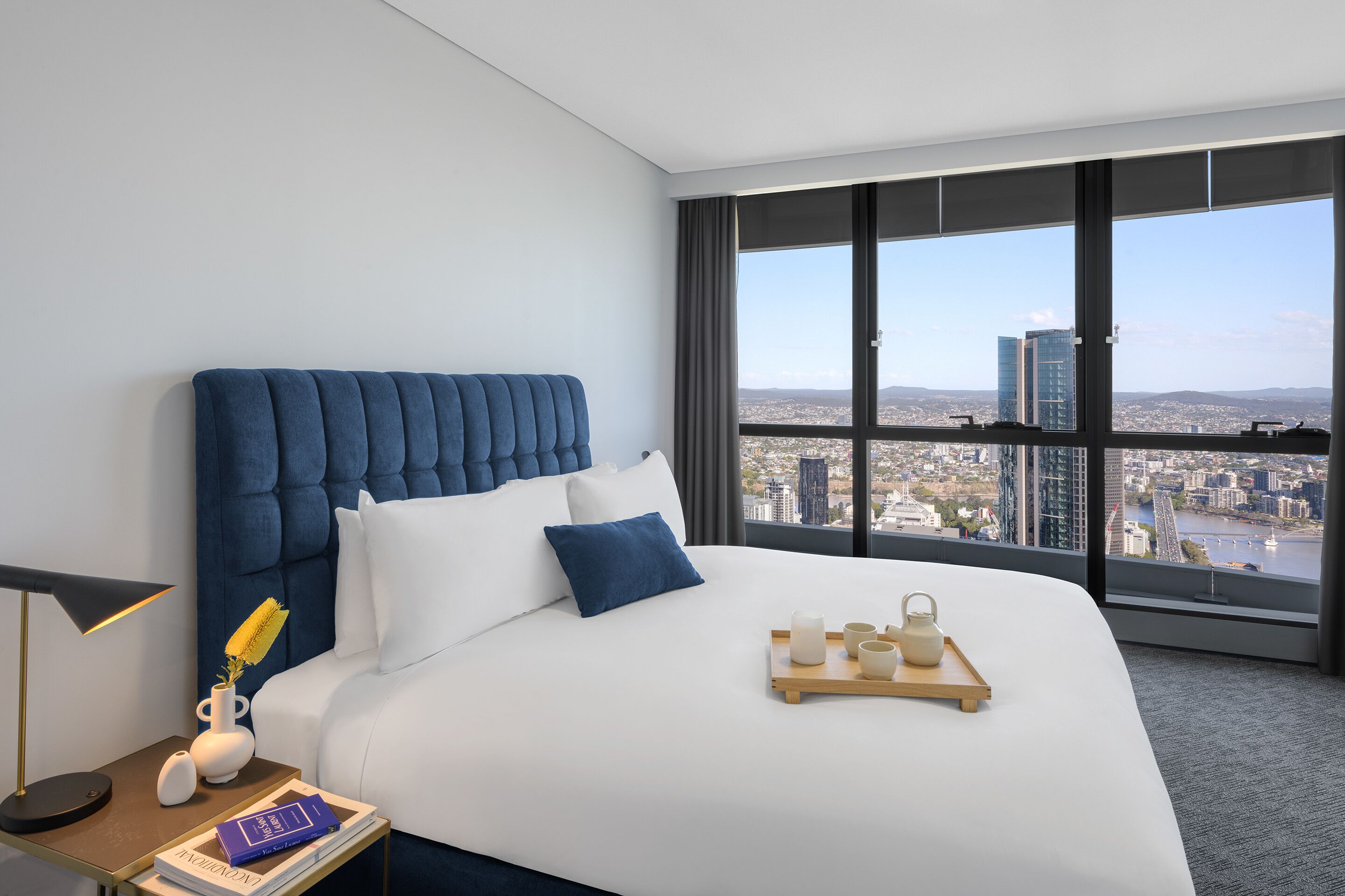 A modern hotel room with a large bed, a nightstand, a lamp, and a view of the city through large windows.