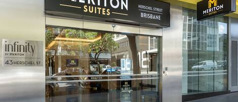 A hotel entrance with a glass facade, signage for 'Meriton Suites' at 43 Herschel St, Brisbane, and a red carpet leading to the entrance.