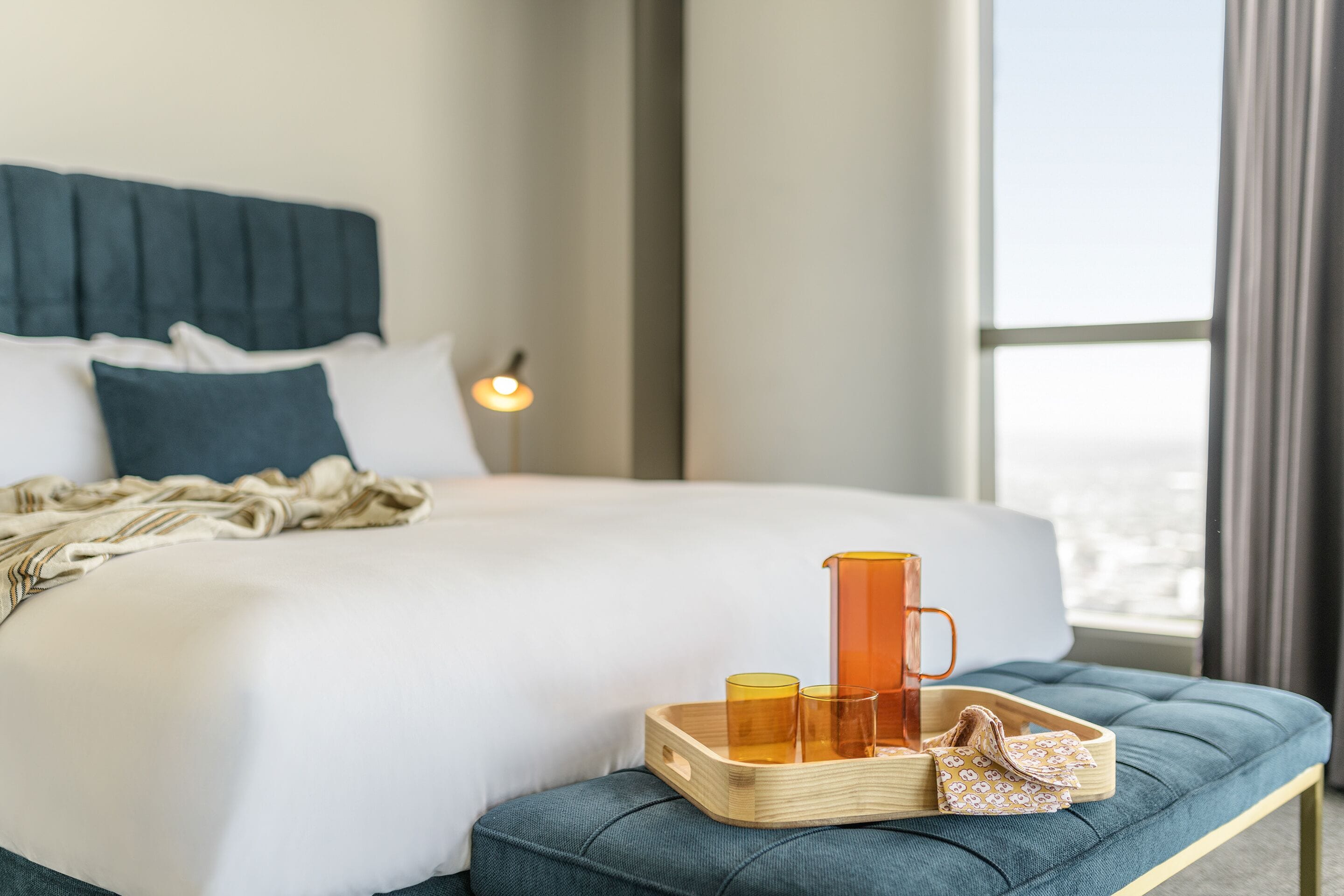 A neatly made bed with pillows, a bedside table with a tray holding glasses and a pitcher, and a view of the outdoors through a window.