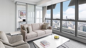 Una sala de estar moderna con un ventanal que ofrece vistas a la ciudad, un sofá color beige, una mesita con una revista y una mesa de centro con fruta.